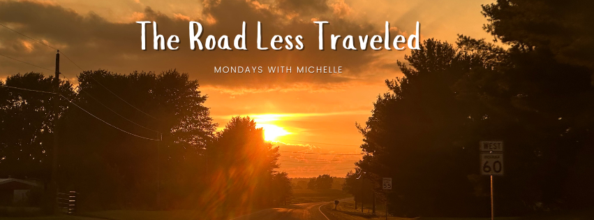 A golden sunset lights up a rural two-lane road flanked by trees, with the sun low on the horizon and a glowing orange sky. A "West US 60" road sign is visible on the right. Overlaid white text reads: "The Road Less Traveled – Mondays with Michelle."
